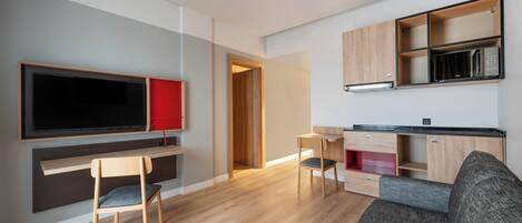 A modern living room with a flat-screen TV, wooden furniture, a grey sofa, and a kitchen area with wooden cabinets and a microwave.