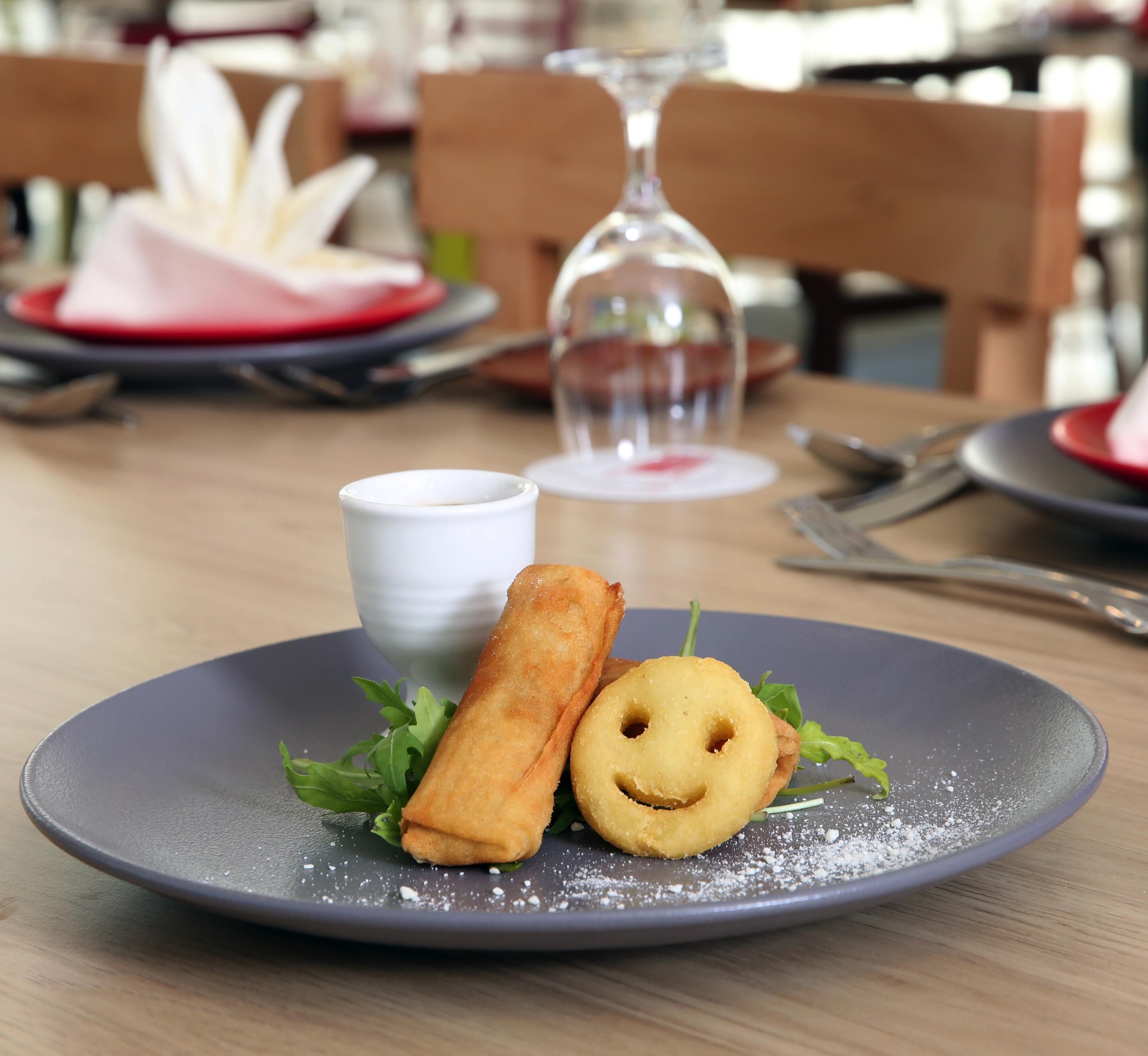 Une assiette avec un plat frit et un biscuit en forme de visage souriant, une petite tasse blanche et un verre d’eau sur une table.