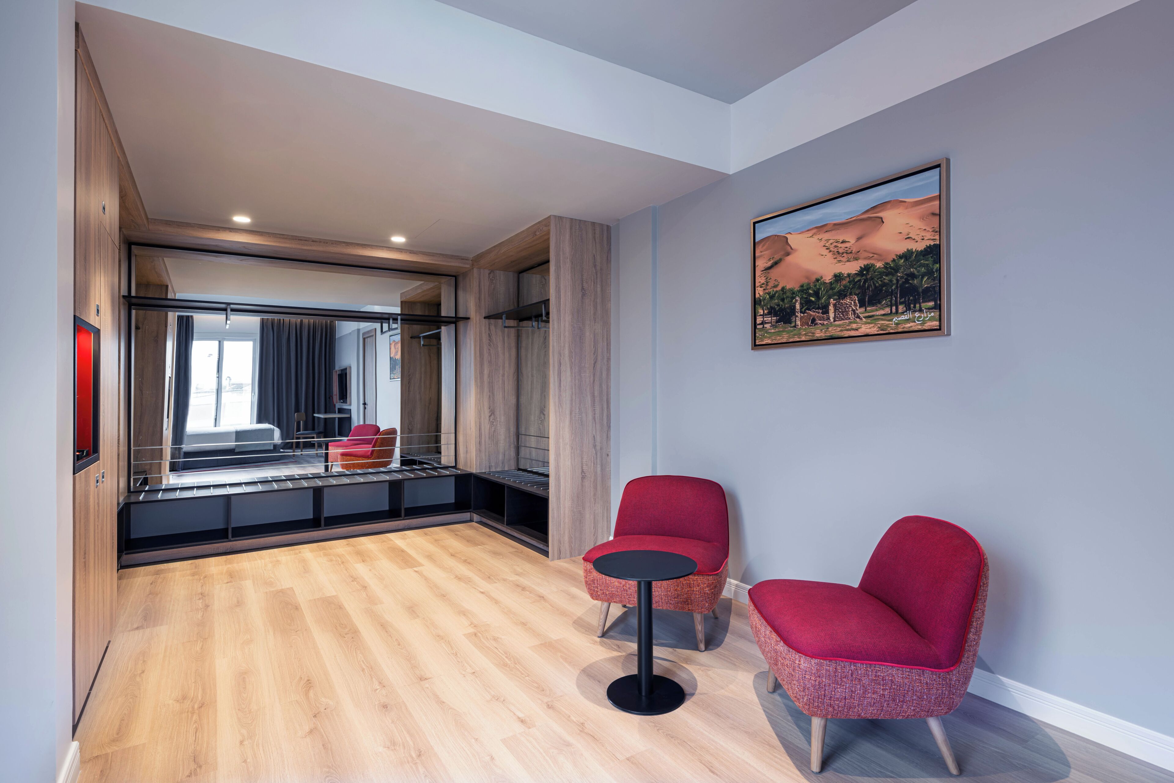 A modern room with a wooden floor, two red chairs, a small table, a large window, and a painting of a desert landscape.