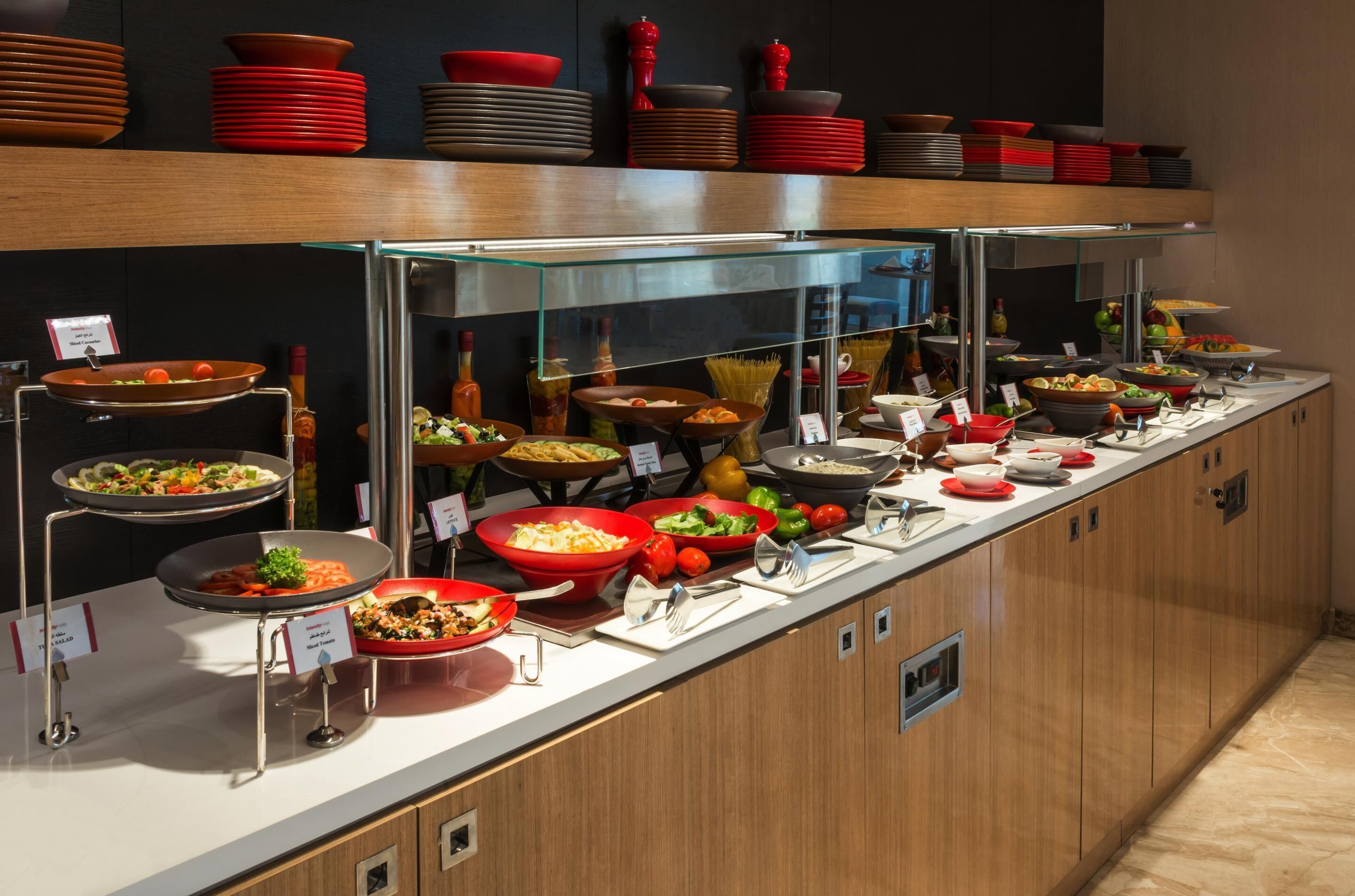 A buffet with various dishes and utensils, a glass display case, and stacked plates on shelves.