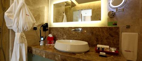A bathroom with a marble countertop, a large mirror with lights, a towel rack, and a robe hanging.