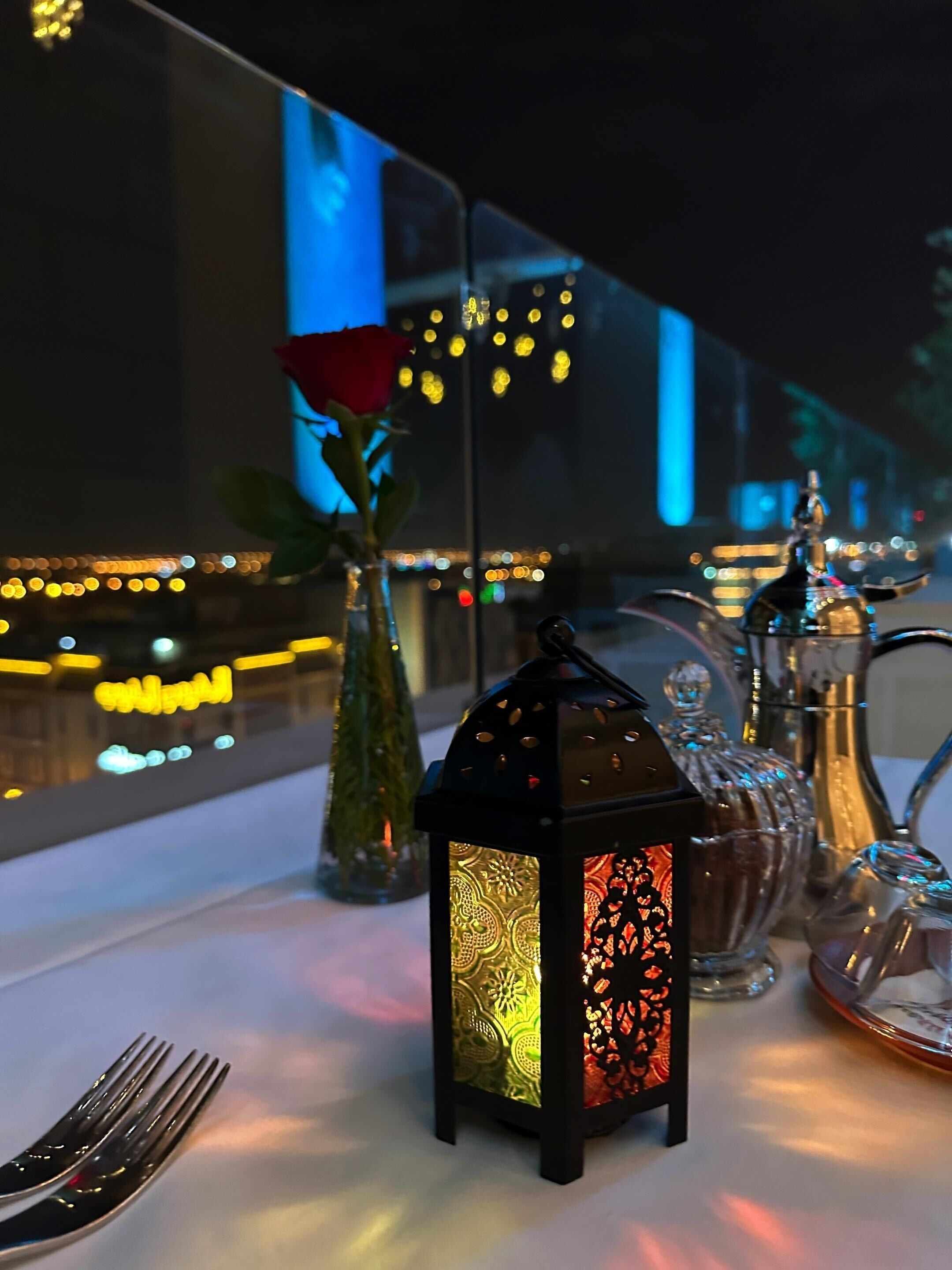 A table with a decorative lantern, a rose in a glass vase, and a silver pitcher.