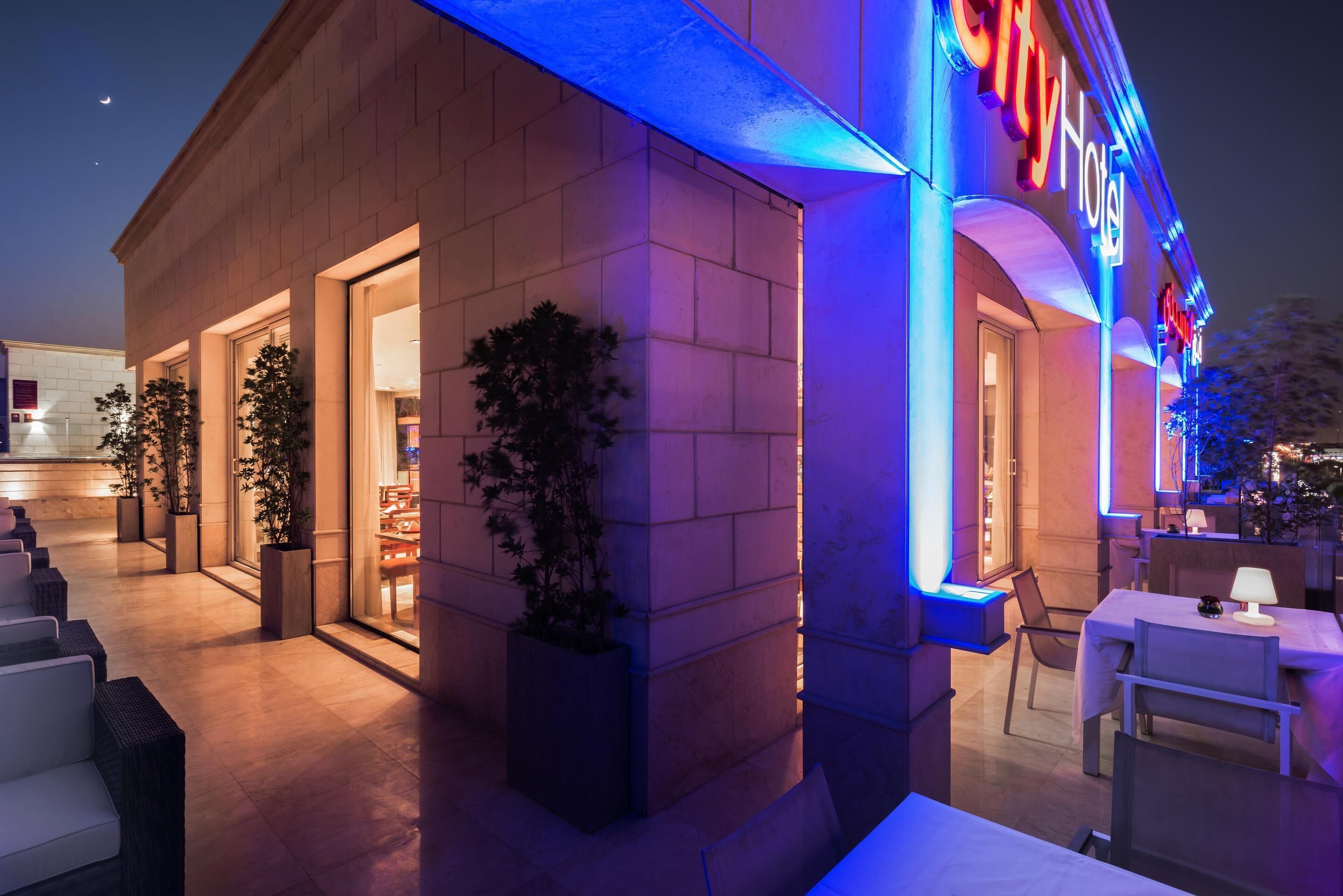 A hotel exterior with a neon sign, outdoor seating, and potted plants.