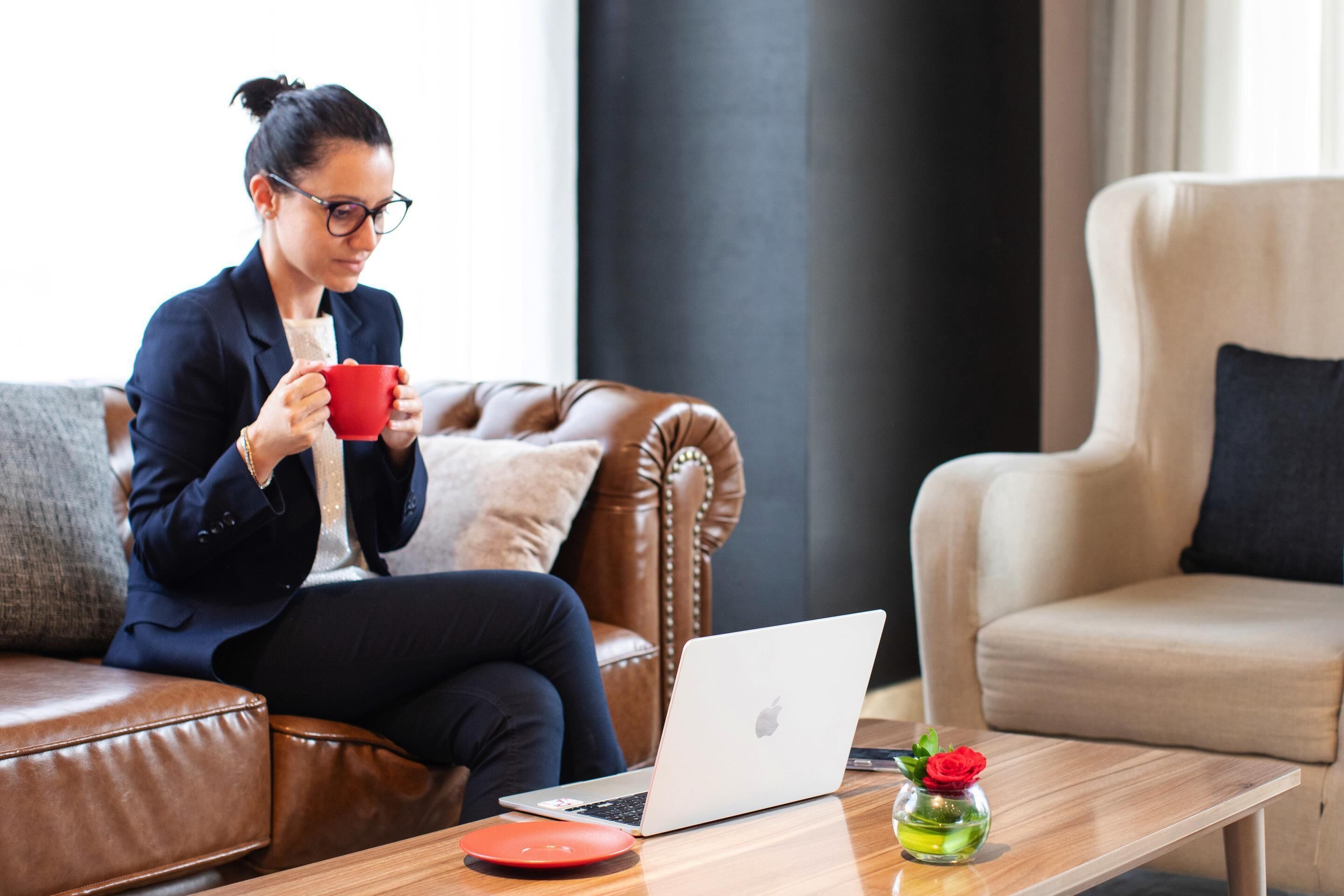 A person sitting on a brown leather sofa with a laptop and a red mug on a wooden table.