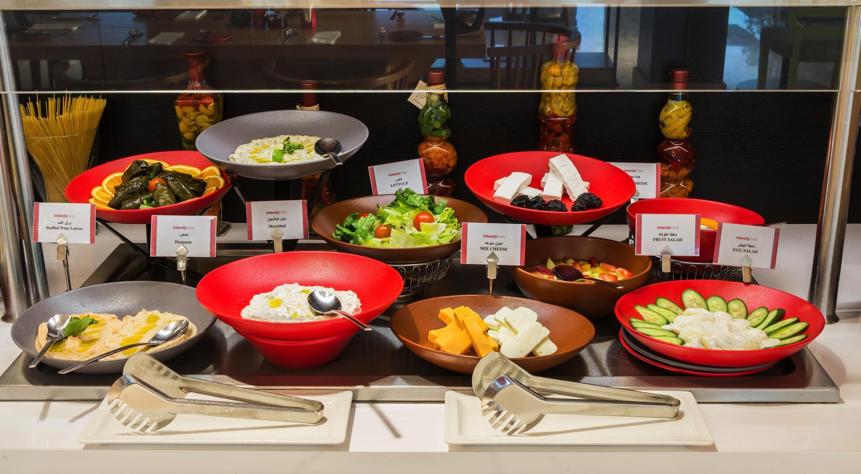 A buffet with labeled dishes including stuffed wine leaves, hummus, mix cheese, fruit salad, and egg salad.