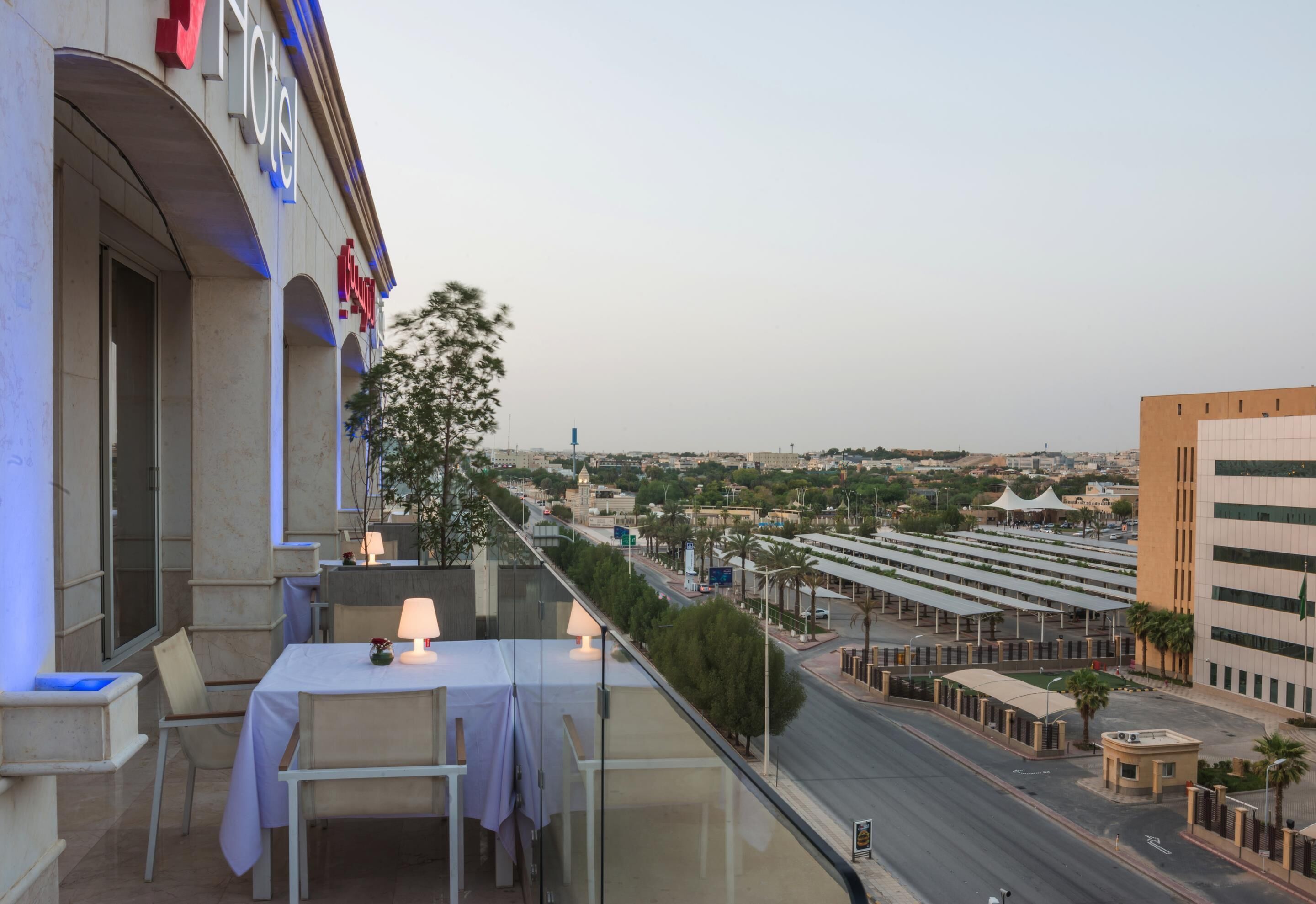 A hotel with a balcony overlooking a cityscape, featuring a table with a lamp, a view of the street, and a building with a sign.