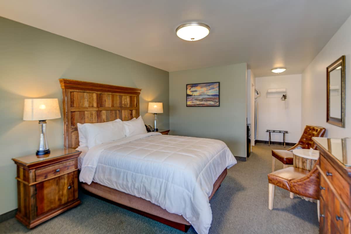 A hotel room with a large bed, wooden headboard, two bedside tables with lamps, a chair, a dresser, and a painting on the wall.