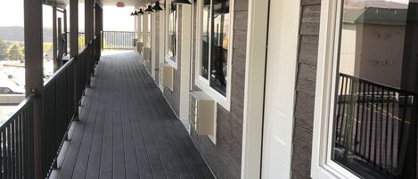 A covered wooden deck with a railing, large windows, and a view of the outdoors.