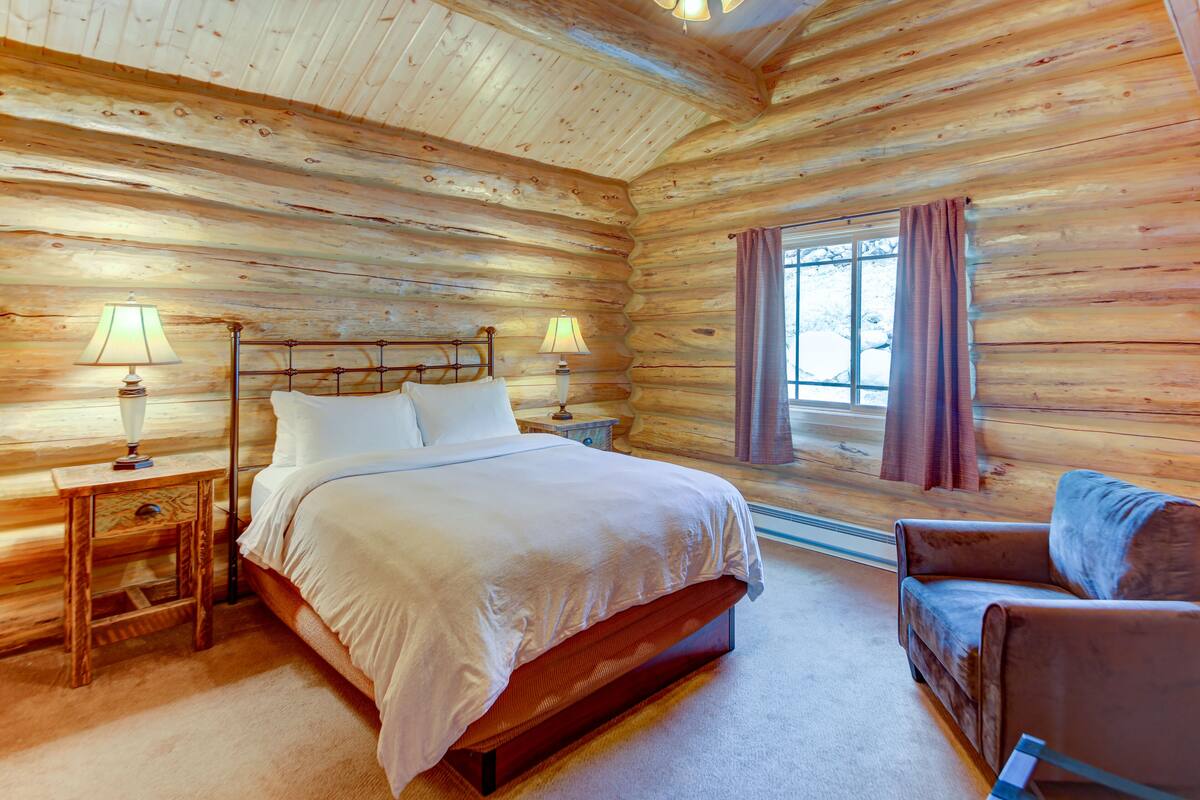 A log cabin bedroom with a wooden bed, two bedside tables with lamps, a window with curtains, and a leather armchair.