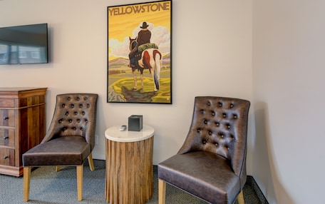A room with two brown tufted chairs, a wooden sideboard, a round wooden table with a remote control, and a wall-mounted television.