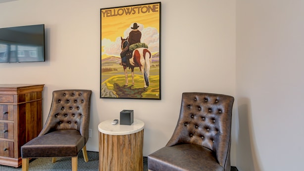 A room with two brown tufted chairs, a wooden sideboard, a round wooden table with a remote control, and a wall-mounted television. - The Roosevelt Hotel - Yellowstone (Gardiner)