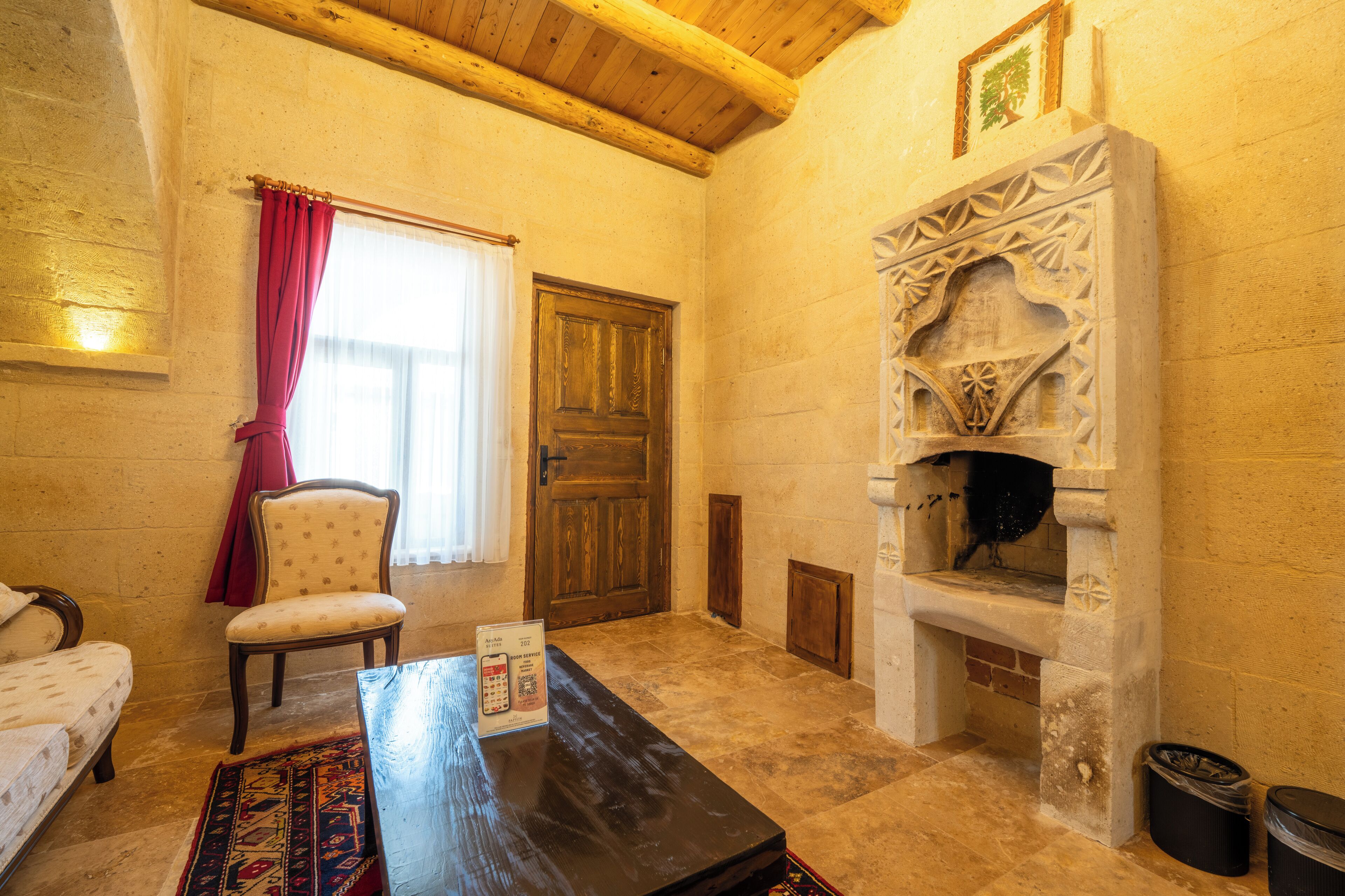 A room with a wooden ceiling, a fireplace, a chair, a table, and a bed.
