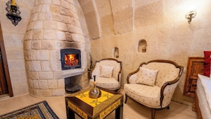 A stone fireplace with a fire, two upholstered chairs, a small table with a decorative vase, and a patterned rug. - Asyada Suites Hotel (Ürgüp)
