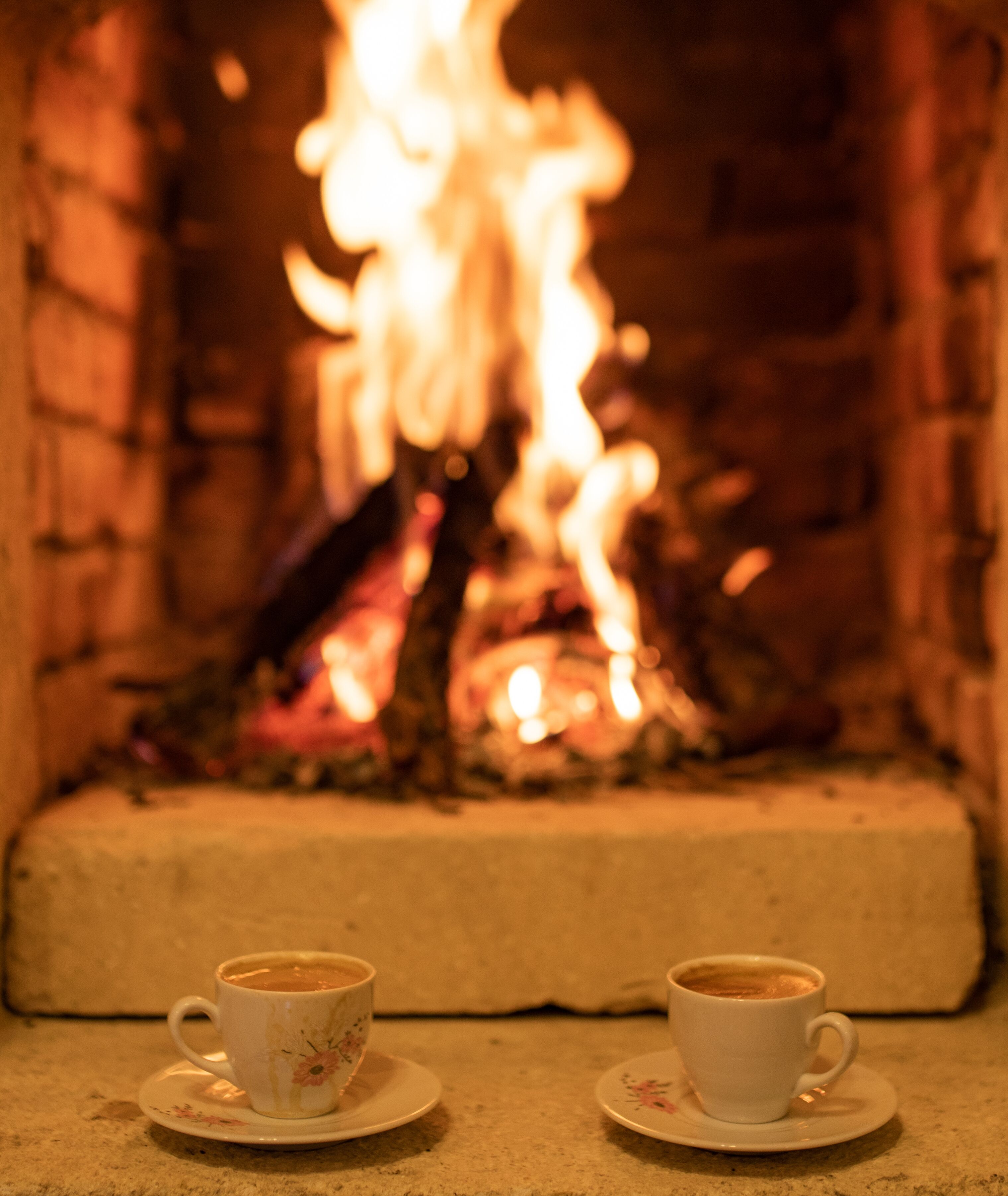 Two cups of coffee on saucers in front of a fireplace with a fire.
