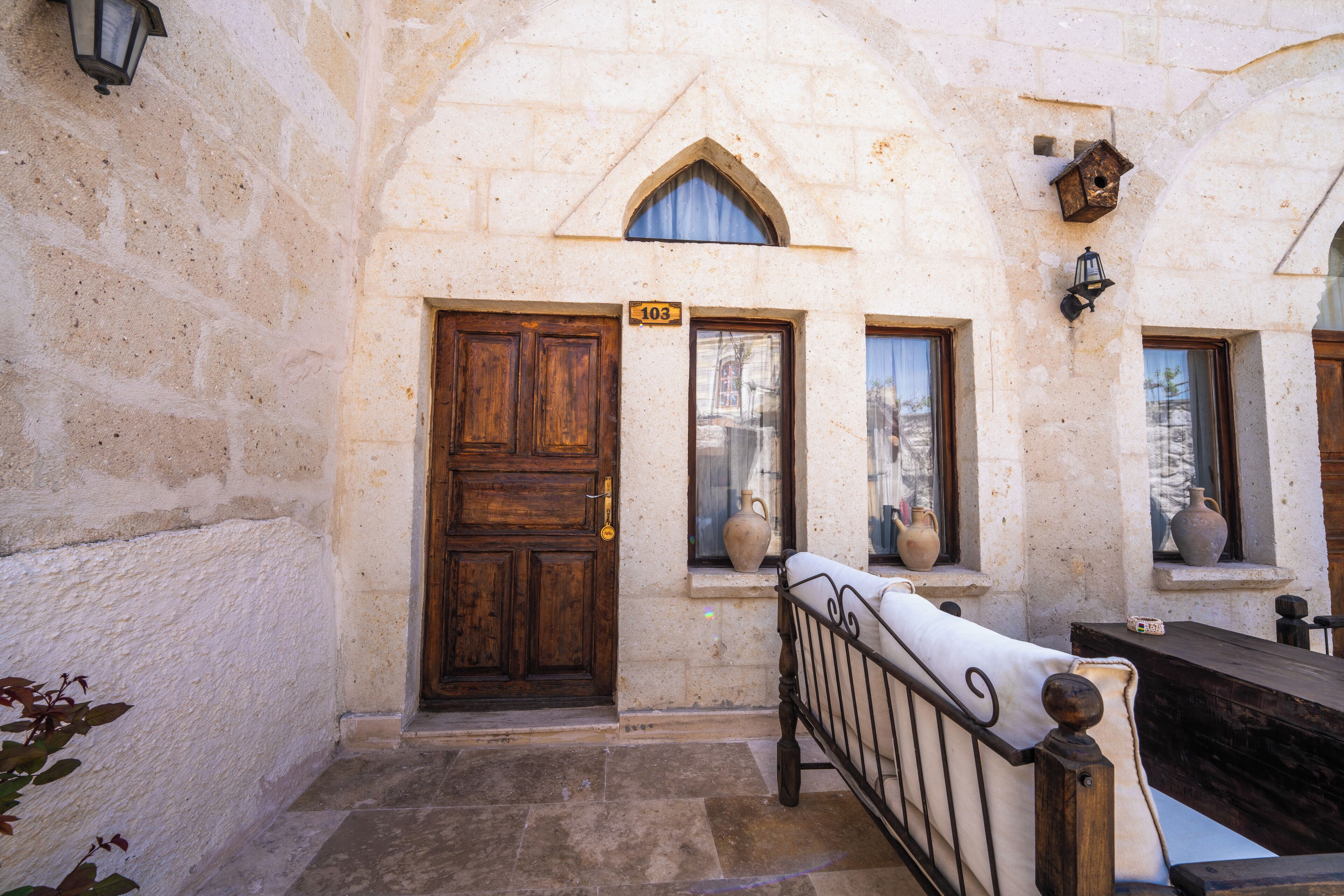 A stone building with a wooden door marked with the number 103, a small window with a vase, and a metal railing in the foreground.