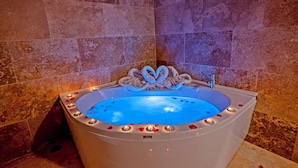 A large, oval-shaped bathtub with a blue interior, surrounded by candles and rose petals, set against a backdrop of reddish-brown stone tiles. - Asyada Suites Hotel (Ürgüp)