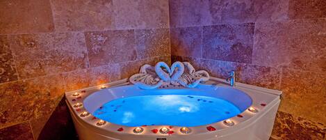 A large, oval-shaped bathtub with a blue interior, surrounded by candles and rose petals, set against a backdrop of reddish-brown stone tiles.