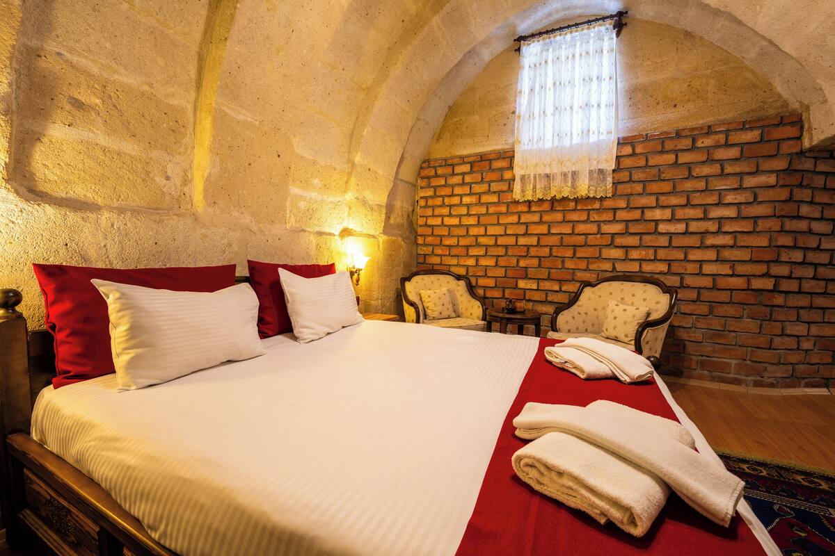 A hotel room with a brick wall, a large bed with white bedding, two armchairs, and a small table.