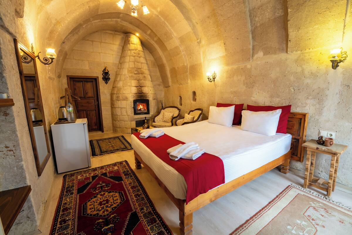 A stone-walled room with a bed, a fireplace, a refrigerator, and a small table with a cup.