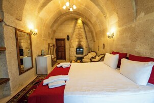Una habitación de hotel con techo de arco de piedra, chimenea, cama con ropa de cama roja, espejo y una mesita con lámpara.