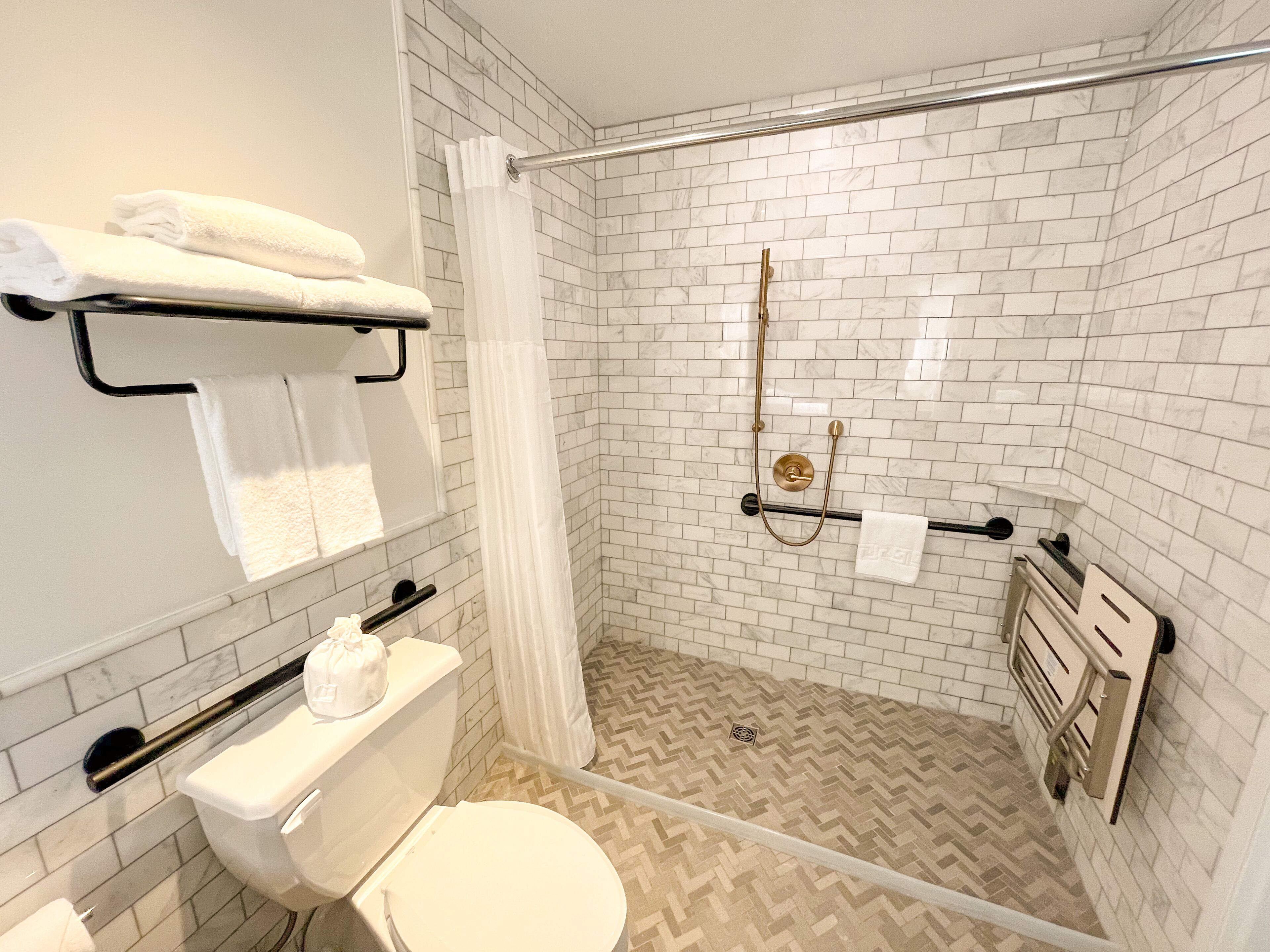 Casa de banho moderna com azulejos brancos tipo metrô, chão em padrão herringbone bege, cabine de chuveiro em vidro e sanita com barra de apoio.