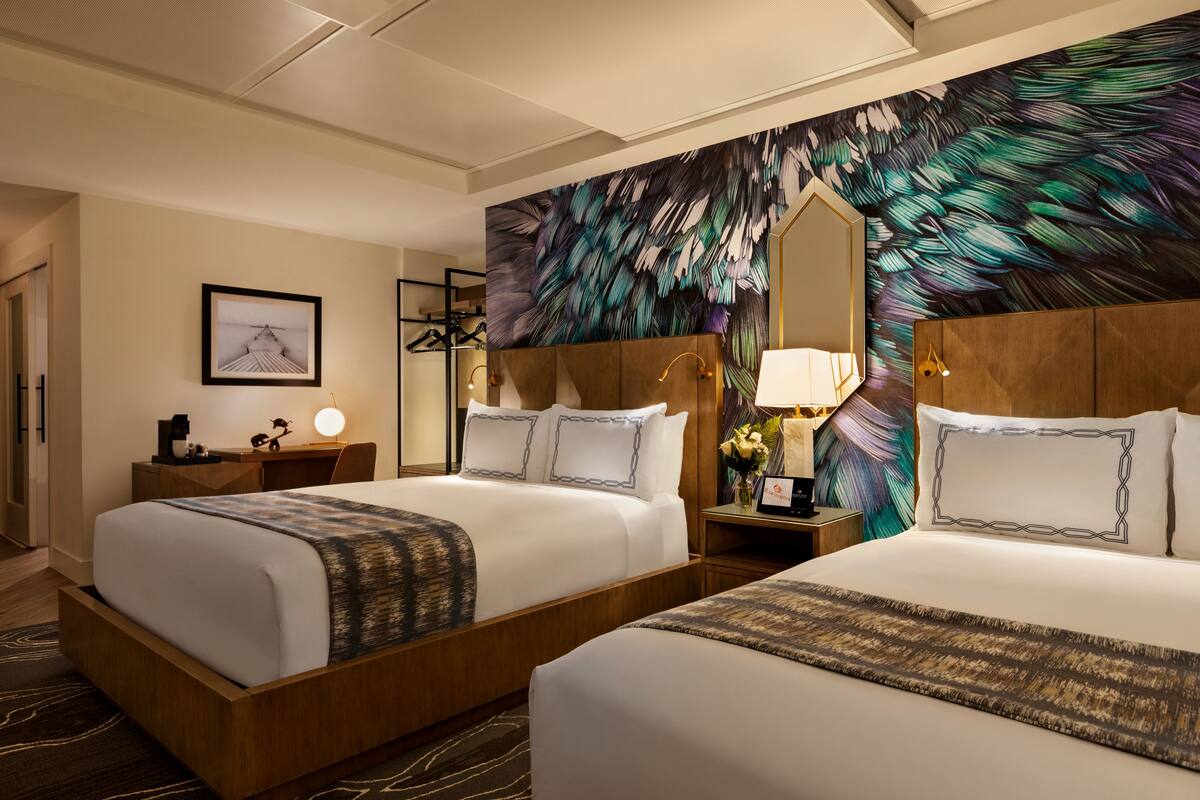 A hotel room with two beds, a large mirror, a nightstand with a lamp, and a wall with a feather pattern.
