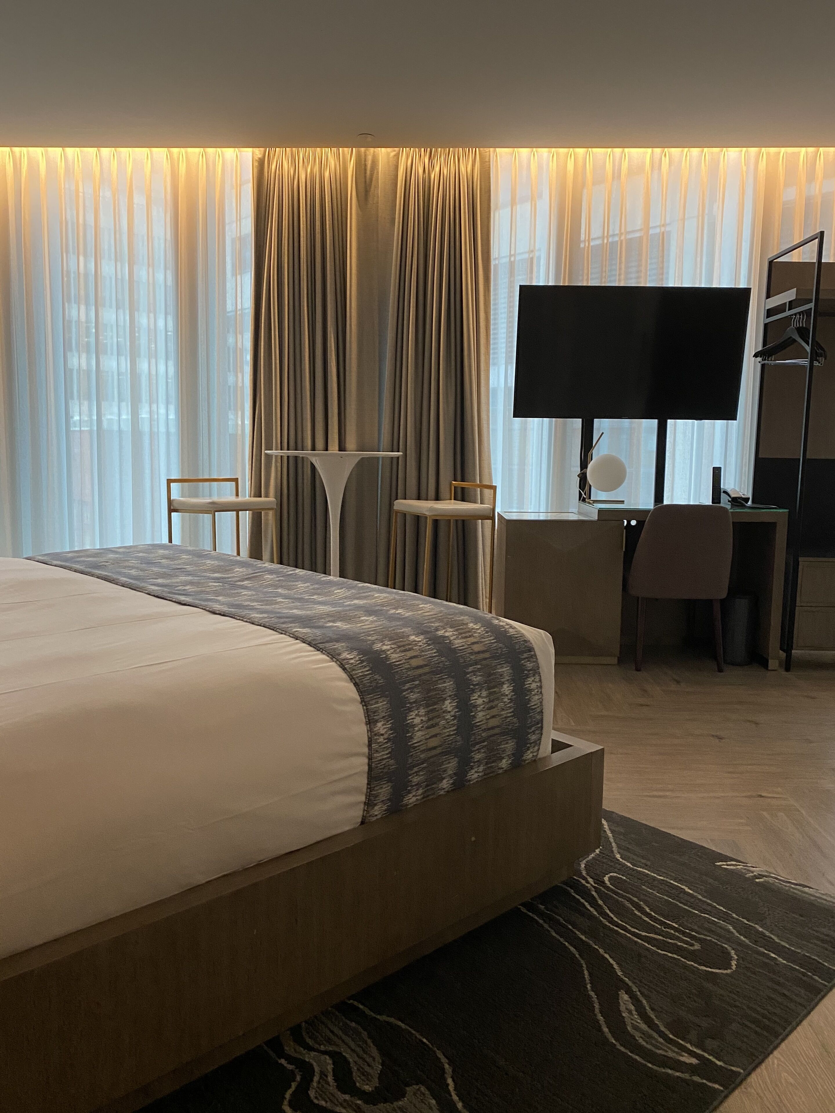 A modern hotel room with a large bed, a desk with a chair, a television, and a wardrobe.