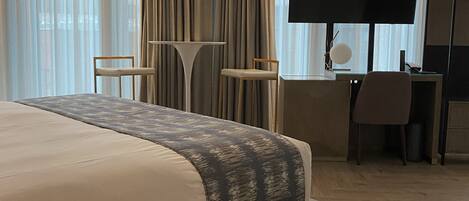 A modern hotel room with a large bed, a desk with a chair, a television, and a wardrobe.
