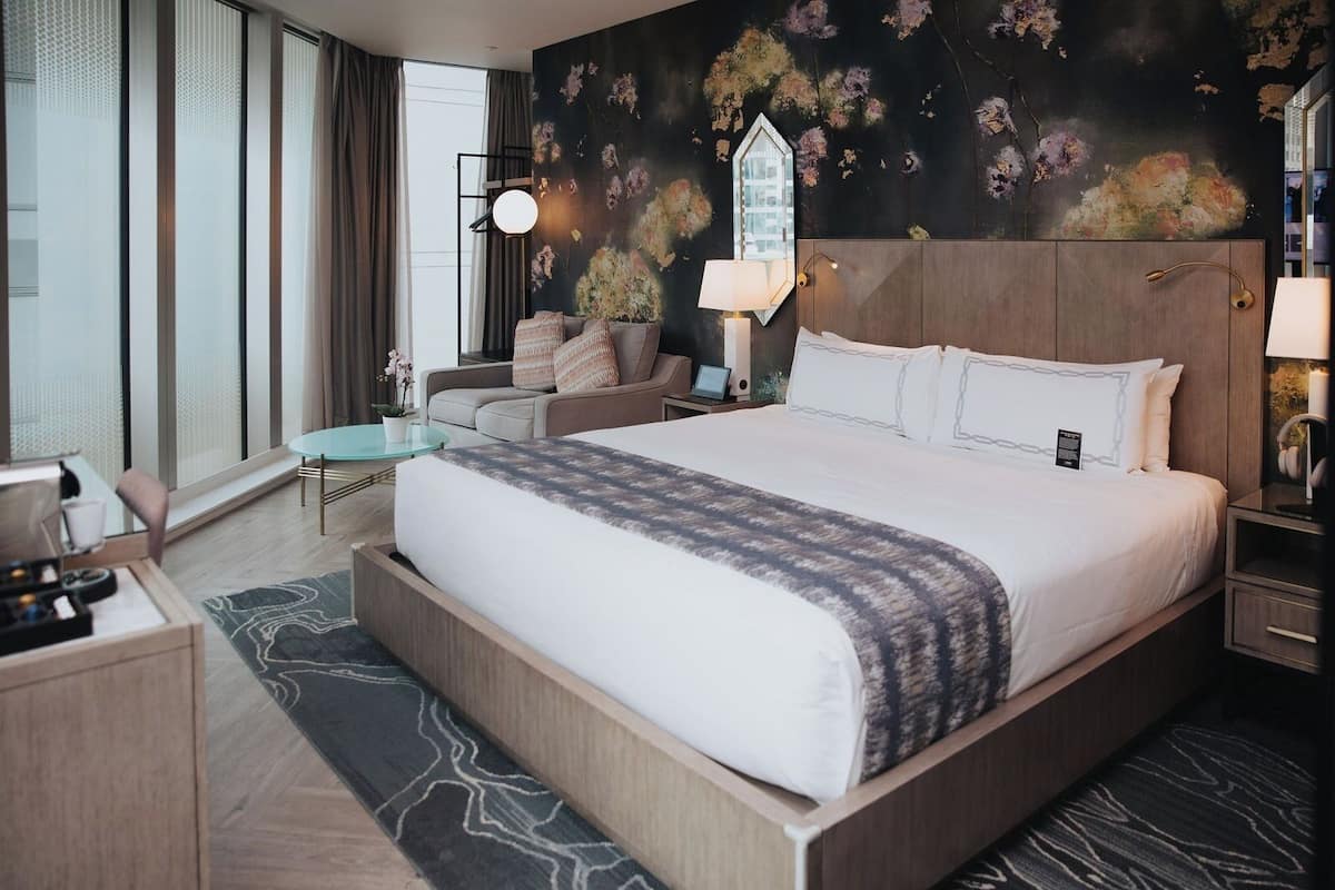 A hotel room with a large bed, a desk, a chair, a small table, and a wall with floral wallpaper.