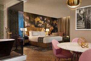 A modern hotel room with a large bed, a round dining table, and a glass-enclosed shower area.