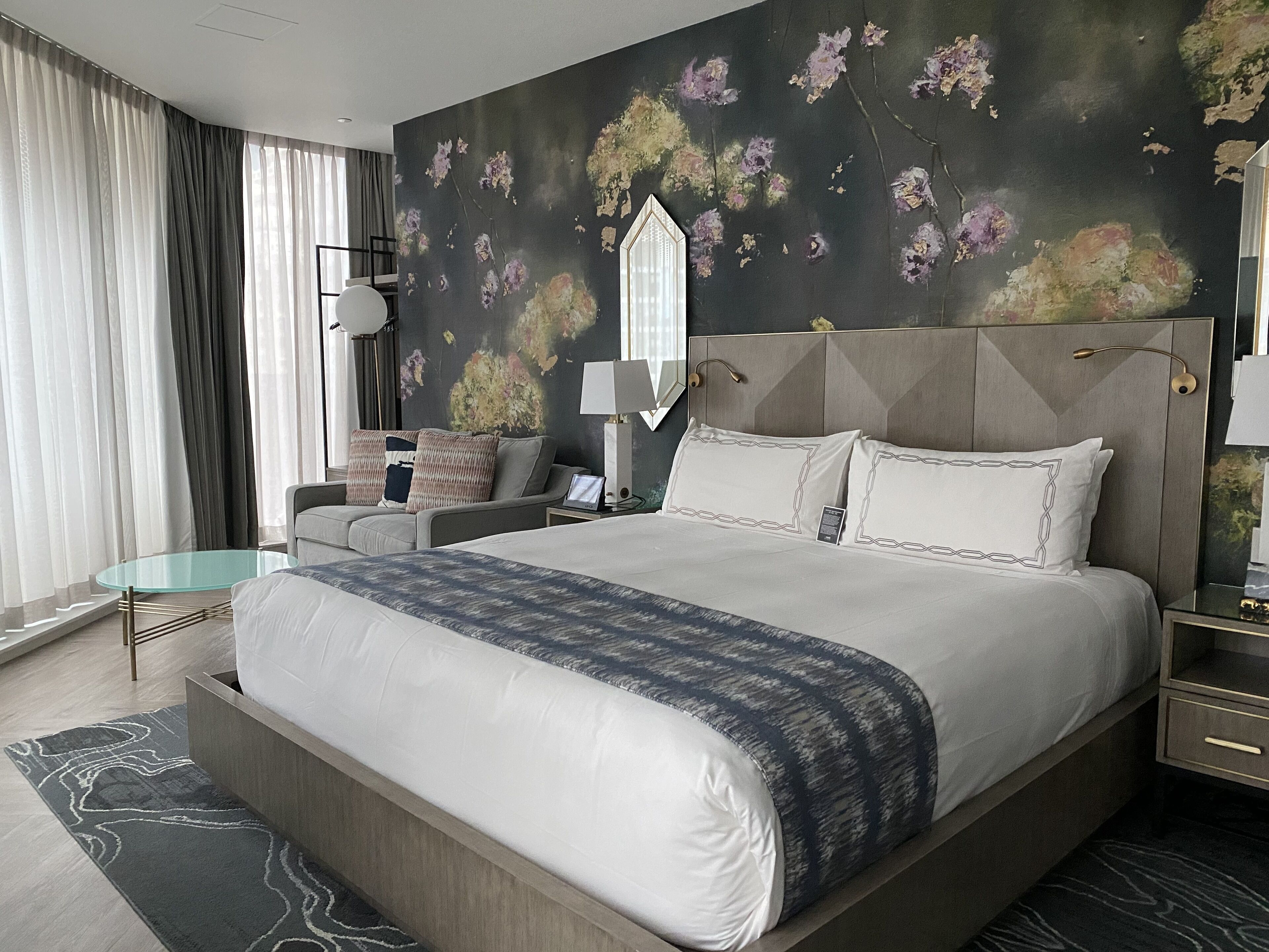 A modern hotel room with a large bed, a sofa, a desk, and a mirror.
