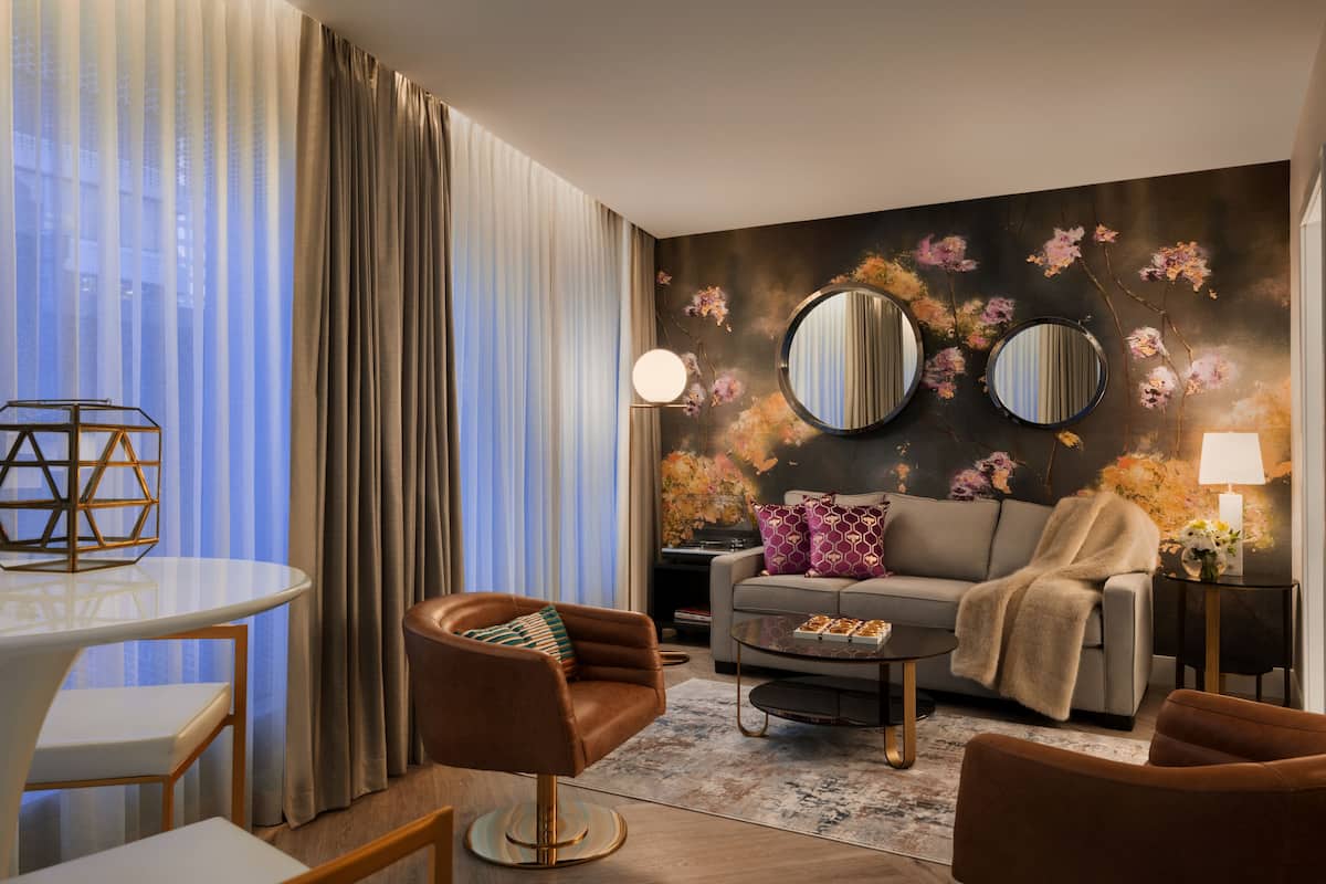 A modern living room with a sofa, armchairs, a coffee table, and a dining table. There are curtains, a lamp, and a wall with floral artwork.
