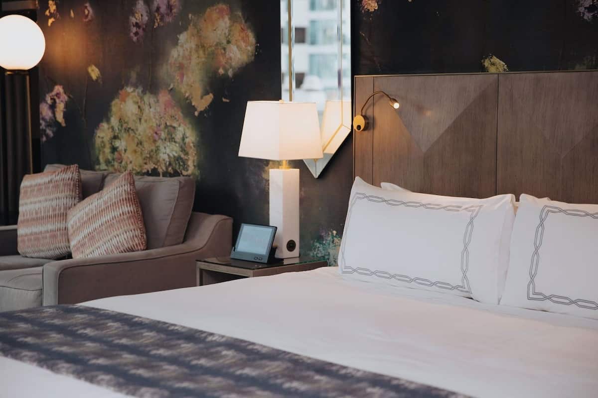 A hotel room with a bed, a desk with a laptop, a lamp, and a couch with pillows.