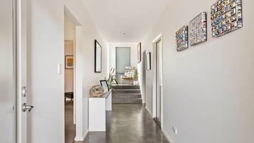 A hallway with a white door, a white console table with a basket, and a chair with a floral pattern. There are steps leading to a room with a large window.