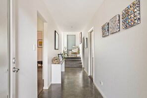A hallway with a white door, a white console table with a basket, and a chair with a floral pattern. There are steps leading to a room with a large window. - Studio 25 Gnarabup - Margaret River - sleeps 4 (Gnarabup)