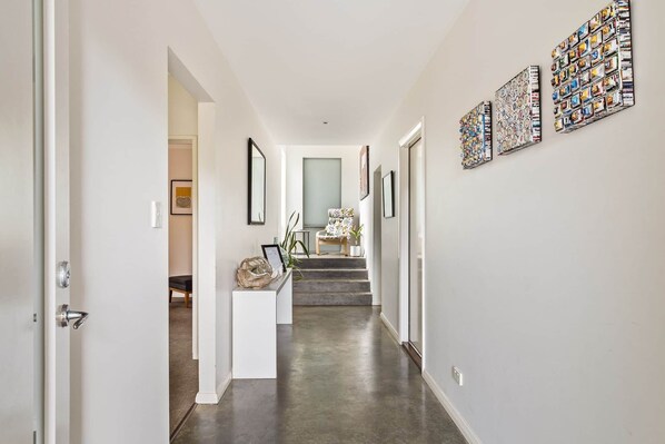 A hallway with a white door, a white console table with a basket, and a chair with a floral pattern. There are steps leading to a room with a large window.