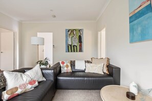 A living room with a black leather sofa, a white armchair, and a coffee table. There is a painting on the wall and a lamp on a side table.