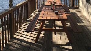 Une terrasse en bois avec des tables de pique-nique, donnant sur un lac, entourée d’arbres et d’un chalet.