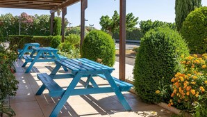 Un espace extérieur abrité, aménagé avec des tables de pique-nique bleues, entouré de haies bien entretenues et de plantes en fleurs.