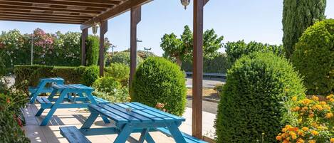 Un espacio de estar al aire libre cubierto, con mesas de picnic azules, rodeado de setos bien cuidados y plantas con flores.