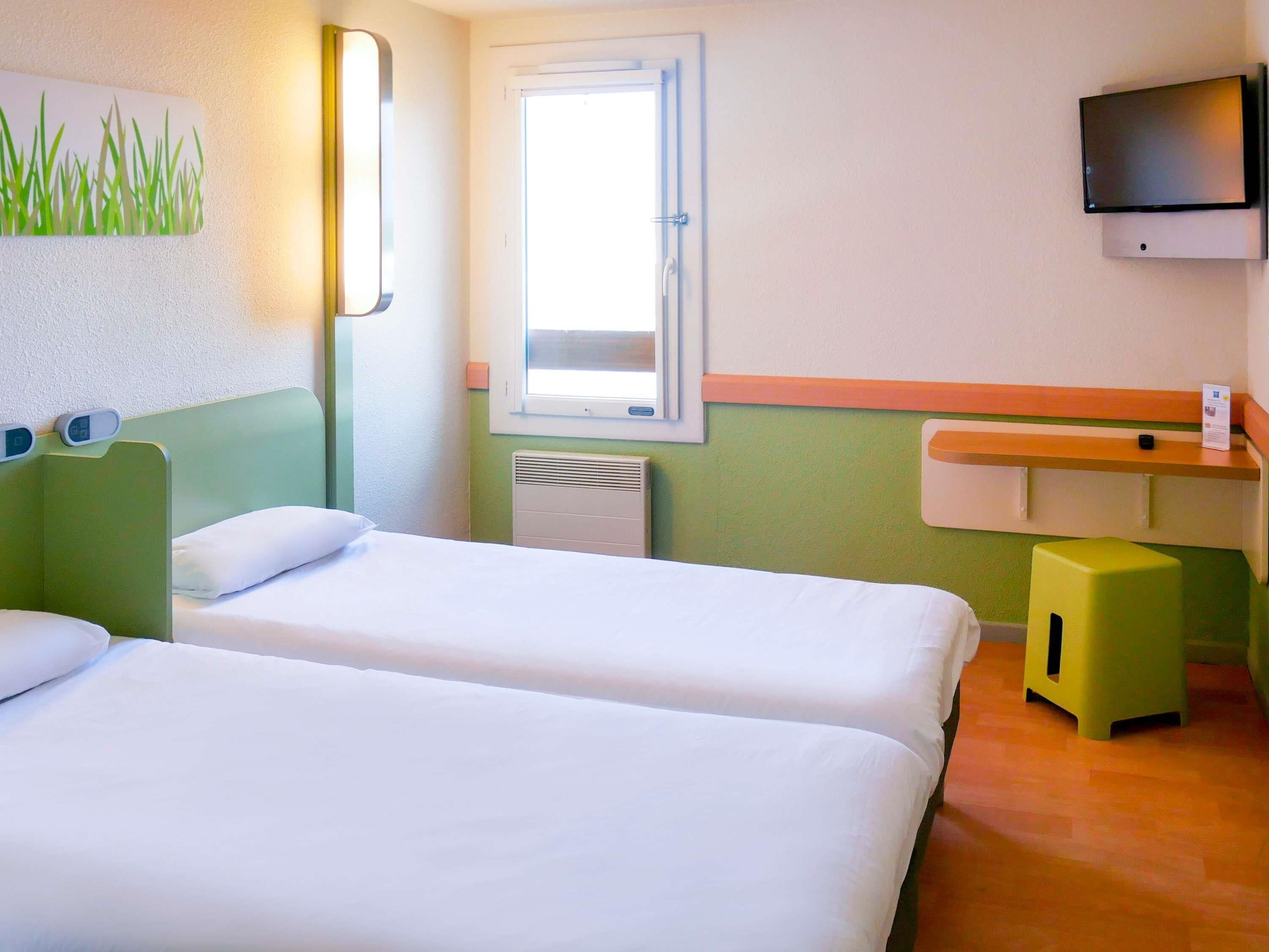 Ein Hotelzimmer mit zwei Einzelbetten, einem Fenster, einem an der Wand befestigten Fernseher und einem kleinen Hocker.