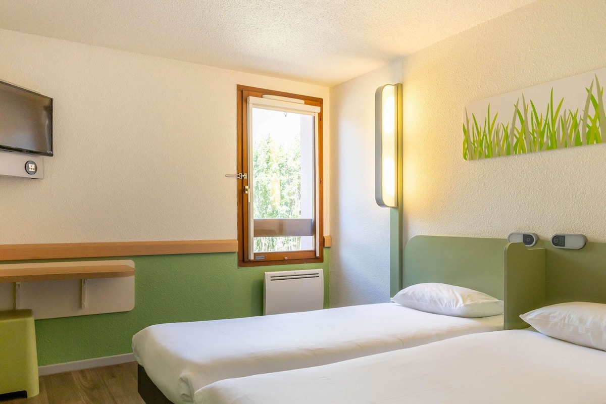Habitación de hotel con dos camas, un escritorio pequeño, un televisor, una ventana y una lámpara fijada a la pared.