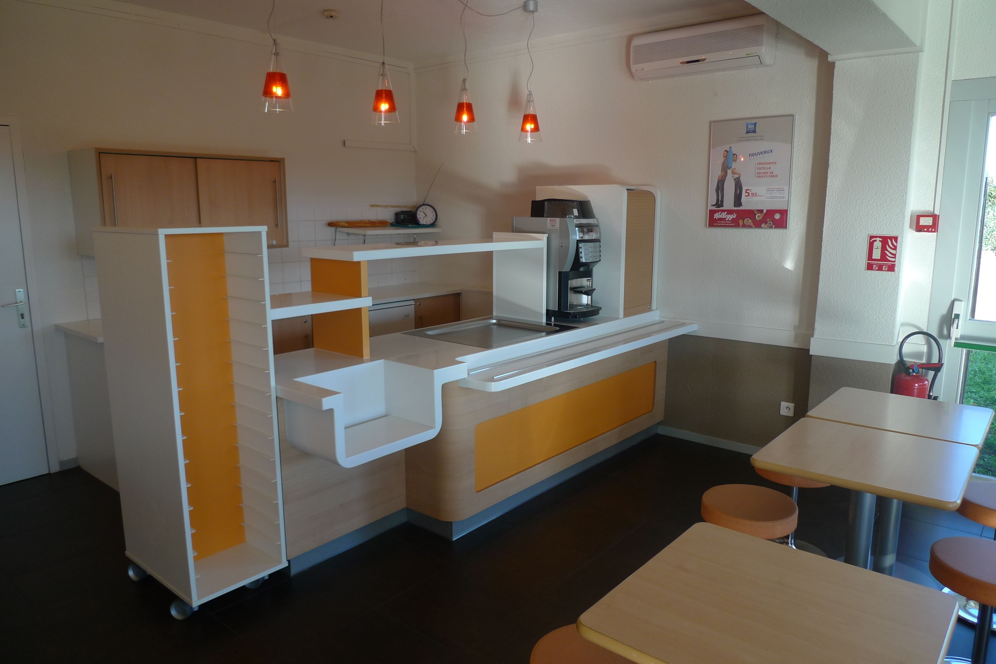 A compact kitchen with a coffee machine, a small refrigerator, and a microwave. There are three pendant lights, a clock, and a fire safety sign on the wall.