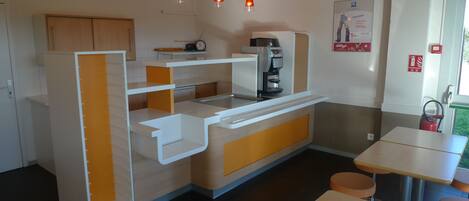 A compact kitchen with a coffee machine, a small refrigerator, and a microwave. There are three pendant lights, a clock, and a fire safety sign on the wall.
