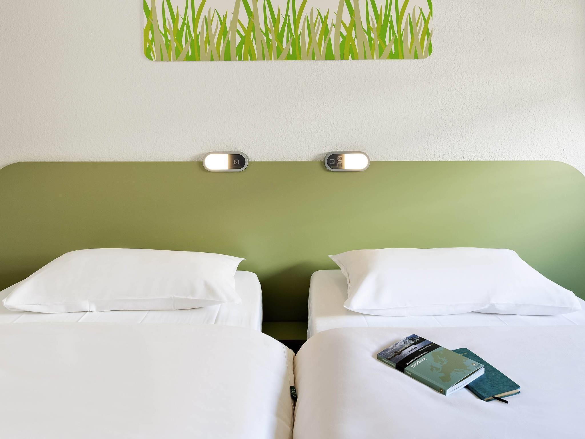 A hotel room with two beds, white bedding, a green headboard, and a wall-mounted light fixture.
