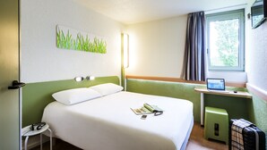 A hotel room with a bed, a small desk with a laptop, a window with curtains, a green stool, and a wall-mounted picture of grass. - ibis budget Caen Nord Mémorial (Saint-Contest)