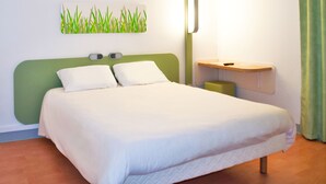 A neatly made bed with white linens and a green headboard, a bedside table with a lamp, and a wall-mounted artwork of grass. - ibis budget Caen Nord Mémorial (Saint-Contest)