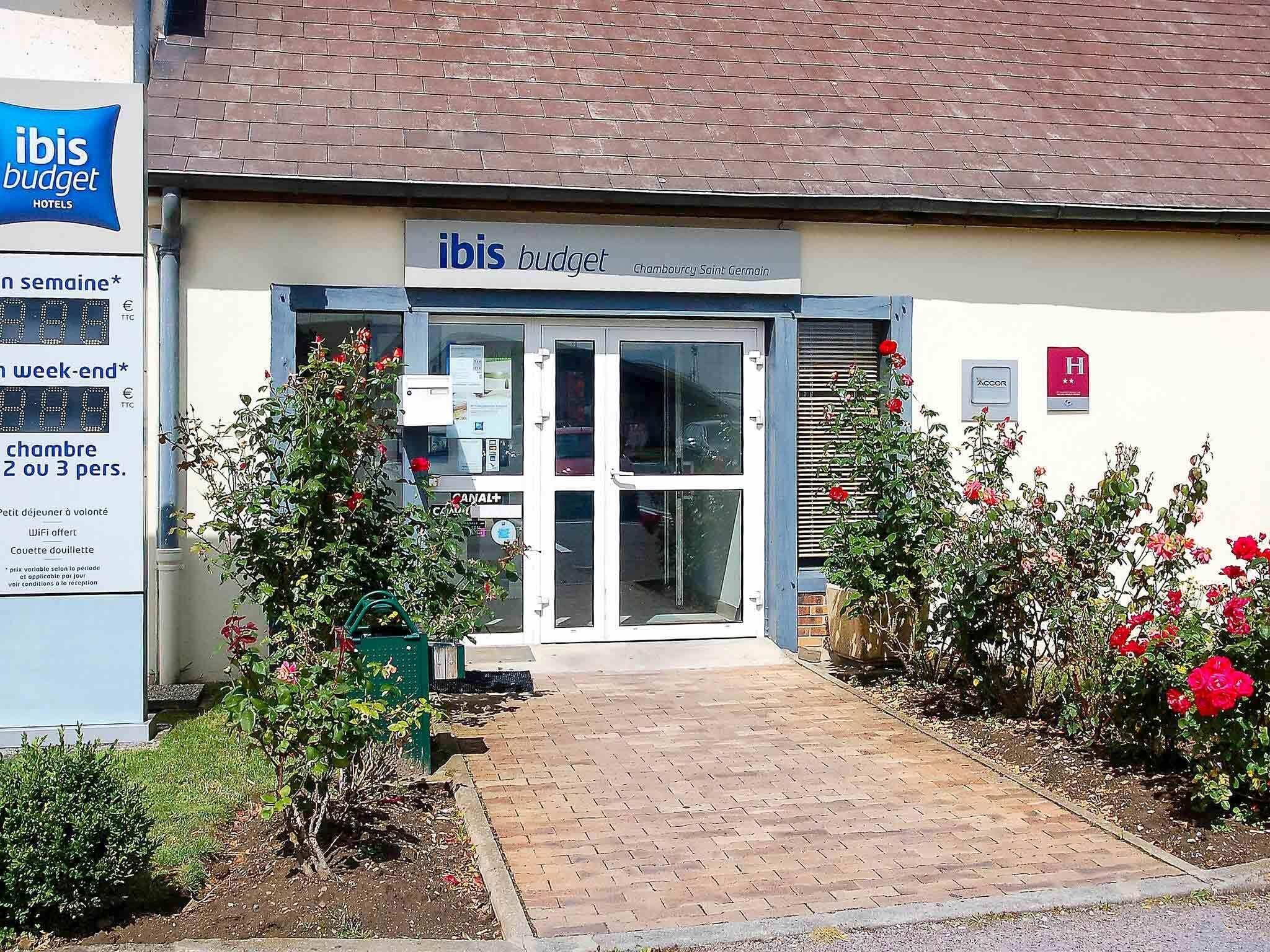 Entrada de hotel con un cartel que dice 'ibis budget Chambourcy Saint Germain', un panel de visualización digital y un pequeño jardín con rosas.