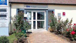 Entrada de hotel con un cartel que dice 'ibis budget Chambourcy Saint Germain', un panel de visualización digital y un pequeño jardín con rosas.
