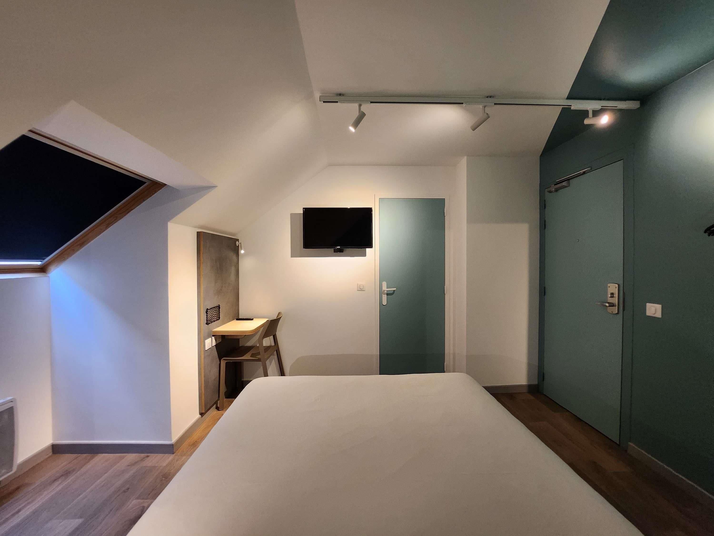 A bedroom with a bed, a desk, a chair, a TV mounted on the wall, and a door.